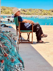 Un hombre contempla el mar turquesa de Menorca sentado junto a las redes de un pequeño puerto pesquero. Un hombre contempla el mar turquesa de Menorca sentado junto a las redes de un pequeño puerto pesquero.