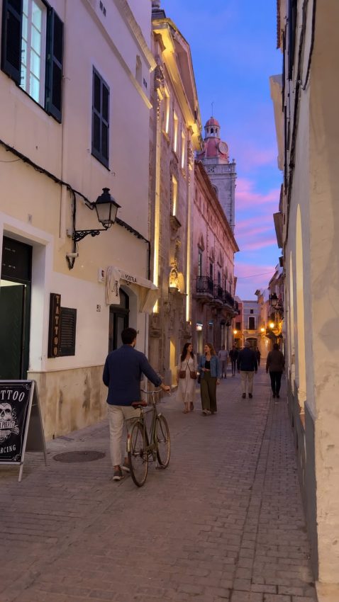 Personas paseando y una bicicleta en una calle de Menorca al anochecer