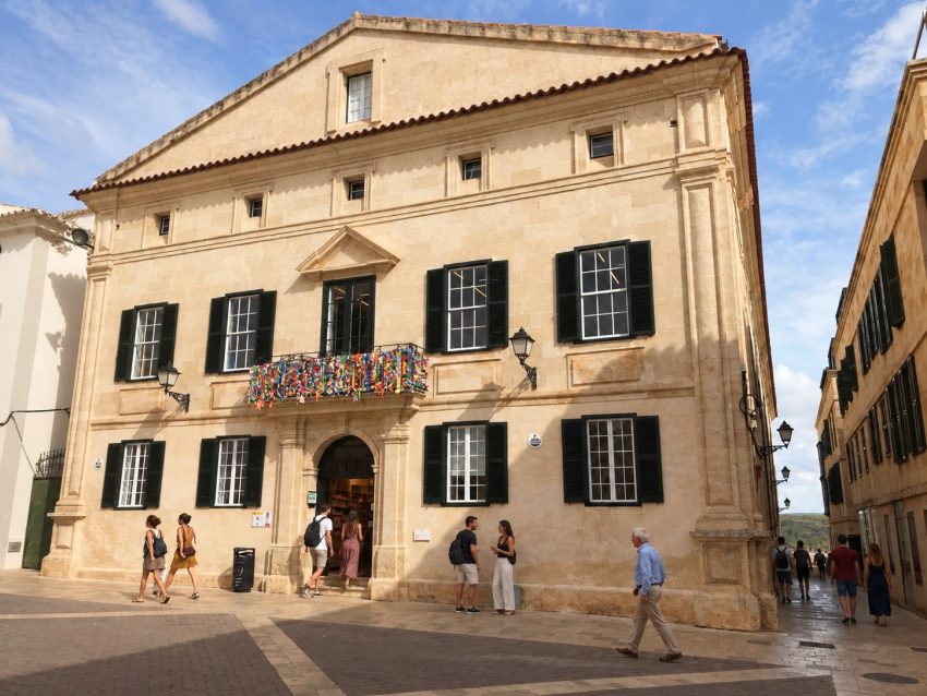 Fachada de piedra con contraventanas verdes y personas paseando en Menorca