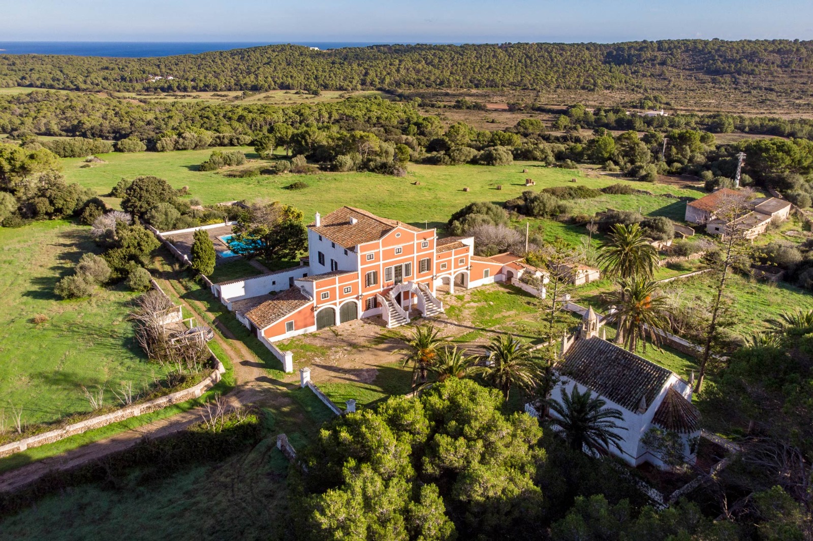 Aerial view of a country house with a terracotta-coloured façade and surrounding land in Menorca